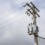 Electricity transformer mounted on a utility pole with overhead power lines against a cloudy sky.